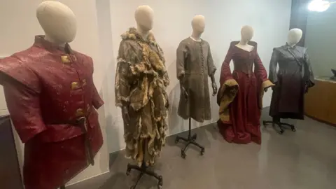 This image shows five mannequins displaying medieval-style costumes from the Game of Thrones series. They include two intricately designed red outfits, one featuring a tunic and the other a long gown with wide sleeves. Other costumes include fur-trimmed attire and long leather coats with chain details.