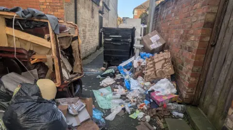 Black bin bags full of rubbish and empty food containers are dumped on the alleyway floor