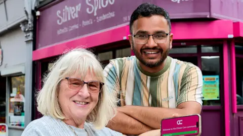 Councillor Maureen Cummings holding a phone showing the Citizen Coin app, pictured with Abrar Nawaz outside his Silver Spice sandwich and curry shop in Wakefield city centre