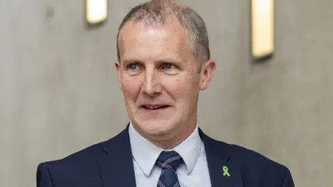 Gret-haired man wearing a navy suit and tartan tie