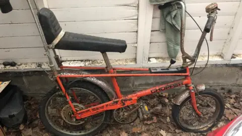 Qays Najm/BBC An orange Raleigh Chopper, with rust on the paintwork and chrome mudguards. It is standing inside a shed with leaves on the floor and white-painted timber walls behind the bike.