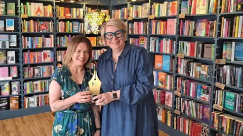 Two women are standing in front of tall shelves filled with books. The woman on the left has brown hair, and is wearing a blue dress. The woman next to her has short blonde hair and blue glasses, with a denim dress on. They are holding a trophy shaped like the nib of a pen