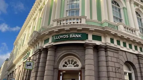 The front of Lloyds Bank on Broad Street in St Helier. In the middle of the image there is a green sign with large white letters which say 'Lloyds Bank'. Above the sign, the building is painted light green, while the pillars are painted white. Below the sign, the building is unpainted brick. To the left of the image, a section of blue sky is visible.