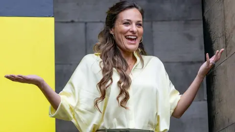 Getty Images Nicola Benedetti in cream shirt stands, laughing and gesturing, in front of a stone building. She has long hair.