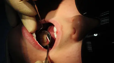 PA Someone receiving dental treatment with instruments in their mouth and wearing a black eye mask.