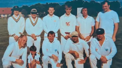 Louise Barrell Ten men wearing cricket whites, and a small boy, in a team line-up on a cricket pitch. Six men are standing in the back row. Four men, and a boy, are crouching kneeling in the front row. Four of the men are wearing cricket caps.