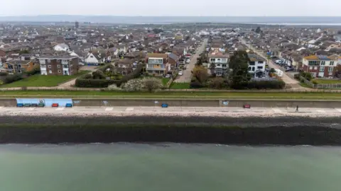 An aerial photo showing the sea in the foreground and rows of houses in the background. It is cloudy.