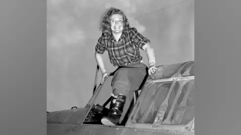 PA Media A black and white photo of Richarda Morrow-Tait standing in the cockpit of a small aeroplane, smiling, her hair blowing in the breeze, in shirt and trousers, her left leg up on the window of the open cockpit, wearing a fur-lined leather boot