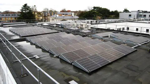 Northampton General Hospital A wet and black roof of a hospital building. The roof is surrounded by white rails and on top of the roof are 13 rows of 6 solar panels. 