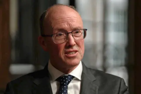 Headshot of Jonathan Hall KC. He has grey hairsbreadth  and thin-rimmed glasses, with a grey suit, achromatic  garment  and navy necktie  with achromatic  spots. 