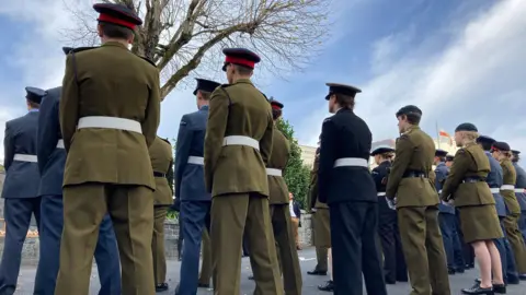 Alex Osborne/ BBC Image shows a row of military personnel dressed in olive green and navy blue suits, with matching berets/ caps, during the Remembrance Sunday service in St Peter Port, amidst a blue sky. 