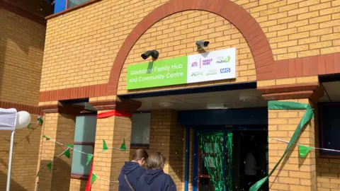 Shariqua Ahmed/BBC Exterior of the family hub building with a green name plate. Two women can be seen walking into the centre, with their backs to the camera.