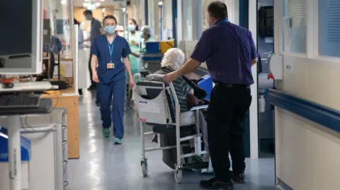 An elderly lady is wheeled along a busy hospital corridor in a wheelchair, with mask-wearing staff present
