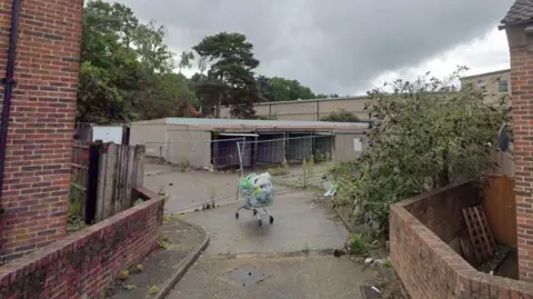 A set of derelict garages which are falling apart. A road leads up to the garages, and an abandoned shopping trolley sits in the middle of the road.