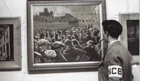 Northumbria University Norman Cornish photographed at the Art by the Miner exhibition in London in 1947. The black and white image shows Cornish pictured from behind looking at a painting portraying a large crowd. Cornish has short black hair and holds a pipe in his mouth. He wears a jacket with a white band around his arm, with white lettering reading WCB.
