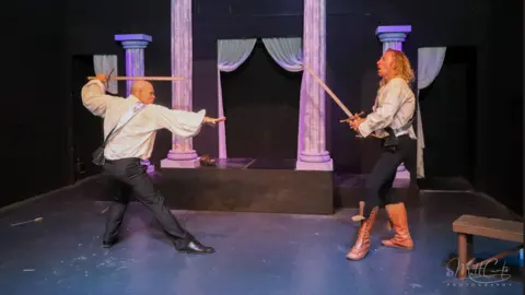 Matt Curtis Photography Two people stand in old style clothes on a stage and brandish swords at each other. There are columns in the background for the set, and everything is lit in purple lighting. 