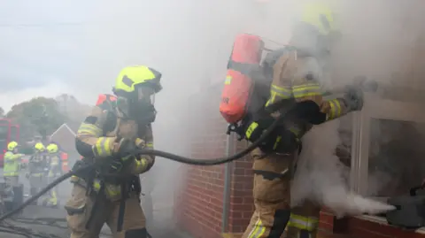 South Yorkshire Fire and Rescue Two firefighters enter a building wearing protective equipment and carrying a hose. A smoke machine is emitting smoke to create a realistic fire environment.