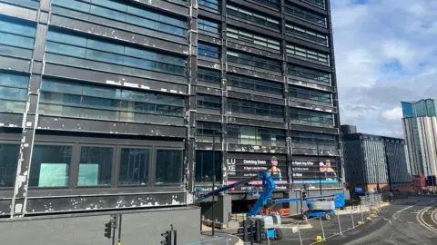 Alexander Brock The exterior of a large building with many windows with a grid-like pattern, and grey steel separating the windows. There are lots of white patches on the steel.