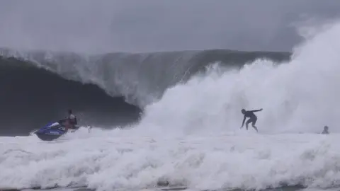 Two men in the sea in stormy weather. One is surfing and the other riding a jet ski.