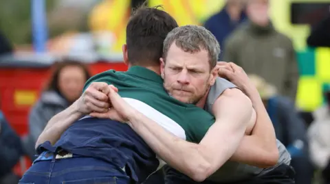 Close-up of two men grappling. One is facing away from the camera. He has dark hair and is wearing a green-and-blue polo shirt. The main facing the camera has dark hair and a beard and is wearing a grey vest. Spectators can be seen in the background.