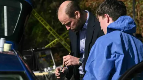 PA Media The Prince of Wales in a dark jacket and blue shirt holds a brush in his hand with a young person in a blue overall looking at him with the side of a car on the left.