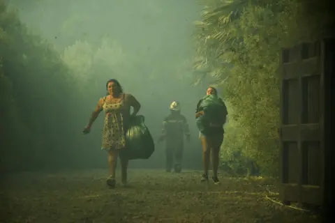 Hector Andrade/Reuters Two women carry their belongings in plastic bags as smoke from wildfire hangs in the air 
