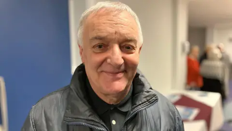 Lou Macari smiles at the camera. He has short white hair and is wearing a black shirt and a black leather jacket.