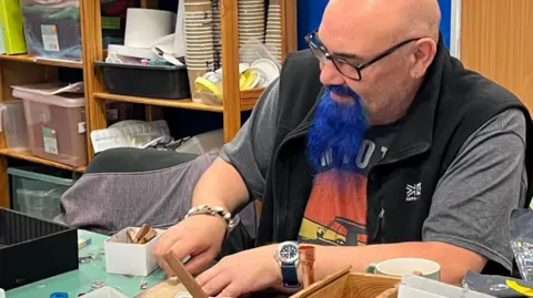 A man at one of the Make and Mend sessions working with wood at a table. He has a bald head and a goatee beard dyed blue. He is wearing black glasses along with a grey T-shirt and black bodywarmer. 