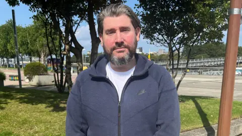 Niall Cahill, a project worker from Glasgow. He has black and grey hair and a beard. He is standing in front of some greenery and his wearing a navy nike jumper and a white t-shirt