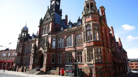A Victorian red‑brick building with a clock tower, red phone boxes, and a post box on a quiet street.
