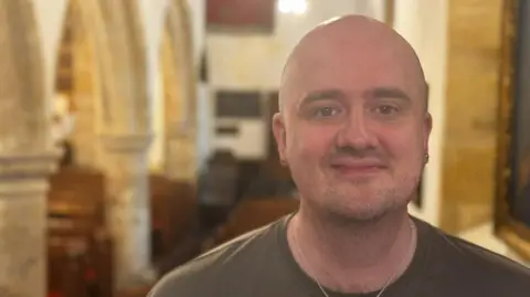 Dominic King/BBC A man stands in a church and looks at a camera.