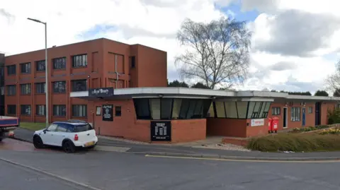 The commercial premises on Well Lane. It is a modern, bricked building with a front entrance. There are cars around. There is a tree in the background and it is overcast.