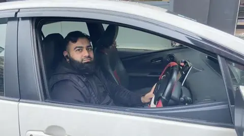 Mohammad Khan sat inside a white car. He has thick black hair and a thick black beard. He wears a black jumper underneath a black body warmer.