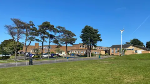 Saint Designs View of the school site at Sheringham. There are multiple brick buildings with trees surrounding them. 