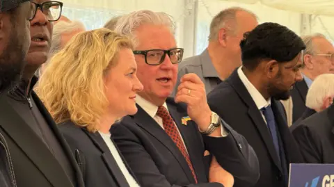 Simon Dedman/BBC Kevin Bentley is in a navy suit and a red patterned tie, wearing glasses with his hair slicked back. He is standing amid a group of people who are smartly dressed, who appear to all be posing for a photo. To his right is Louise McKinlay, who is a blonde-haired woman.