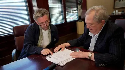 BBC/Wall to Wall Media/Aled Jenkins A meeting takes place around a polished wooden boardroom table, where Sheen and Stewart sit side by side reviewing a printed report. Stewart, who has grey hair and is wearing a pinstripe suit points to text on the top page while Sheen rests a hand on the document. Sheen is wearing a navy overshirt over a green shirt and white T-shirt. Large windows with partially closed blinds line the wall behind them, and office furnishings and artwork can be seen in the background.
