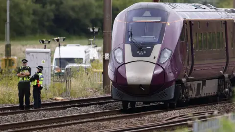EPA/Shutterstock Two police officers, by the side of a purple train, that is on a railway track. There are lights and vehicles to the left. You can see several lines of railway track. 
