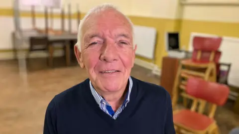 An older male  with thinning white hair is centre of the photo wearing a navy blue jumper and the white and blue striped collar of his shirt is also visible. Behind him and out of focus are mustard and cream colour walls, some wooden red chair are also stacked over his left shoulder and there are some tables stacked in the background over his right shoulder.