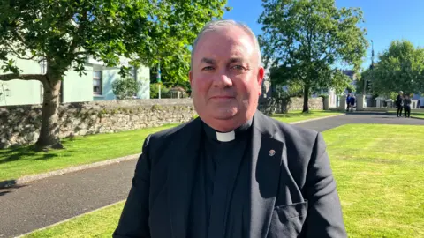 A priest stands in grounds of St Mary's Church in Buncrana looking at camera on sunny day ahead of funeral service for teenage boy Emmanuel Familola who died along with another teenager after getting into difficulties on the County Donegal coast.