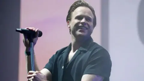 Olly Murs wearing a black button-down top and holding a mic