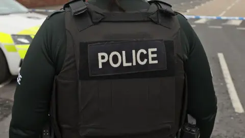 PA Media A close-up image of a police officer's black protection vest as they stand near a cordoned off street.