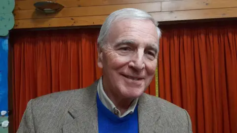 Local Democracy Reporting Service A head-and-shoulders image of Tim Willbond wearing a blue sweatshirt and a grey blazer. He is standing in a hall in front of a red curtain. He is looking at the camera and smiling.