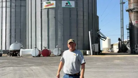 Brad Smith Brad Smith, wearing a grey T-shirt and a baseball cap stands on pavement in the sun, in front of a large cylindrical container.