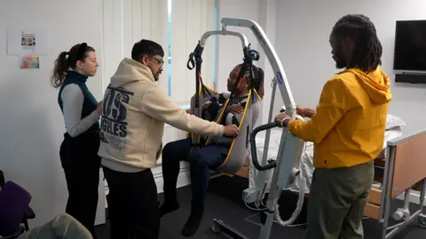 JAMIE NIBLOCK/BBC Four new staff are engaged in hoist training. Two of the staff are standing to the left of the mobile hoist and one of the carers, who is wearing a yellow hoodie, is standing to the right and operating the controls. A female staff member is sitting in the hoist. She has her arms crossed across her chest and a male member of staff is supporting the hoist sling in front of her.