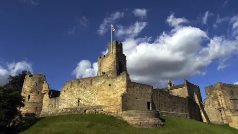 Prudhoe Castle which is large with turrets and slightly damaged. There is a chunk of the stone wall missing from the left hand side. There is a flag pole on the central and highest turret with the red and white flag of English Heritage. The castle sits on a grassy hill and the sky above it is blue with a large white cloud.