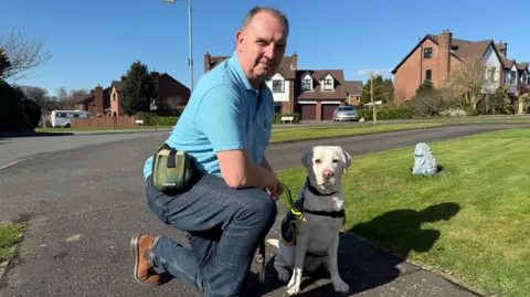 Peter Lockhart. He is wearing a blue polo shirt, jeans and brown boots, he is knelt down next to a white Labrador. Houses and gardens are behind them.