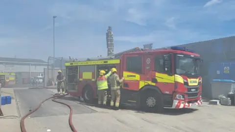 Firefighters are surrounded by white smoke as they direct their hoses towards mounds of rubbish piled up in a concrete bay.