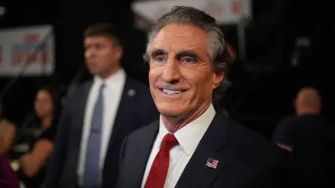 Getty Images Doug Burgum, with salt and pepper hair, and wearing a black suit with a red tie, smiles for the cameras