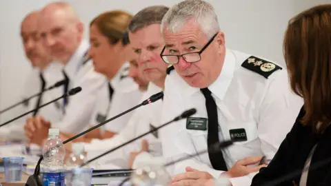 PA Media Jon Boucher leaning over a table speaking into a microphone. Several other senior police officers are sat next to him.