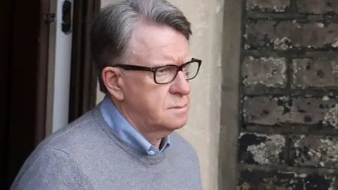 Peter Mandelson, wearing a blue shirt beneath a grey jumper, looking serious as he exits his home.
